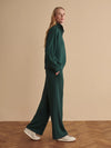 A woman modeling a dark green two-piece lounge set consisting of a long-sleeve mock-neck top and matching wide-leg trousers, paired with white sneakers, set against a warm beige studio background with a wooden floor.