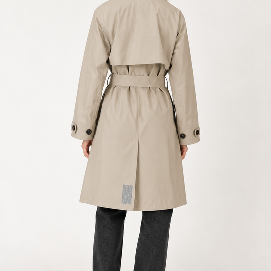 A woman shown from the back wearing a beige trench coat with a belted waist, paired with dark gray jeans and black shoes, against a plain white studio background.