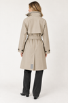 A woman shown from the back wearing a beige trench coat with a belted waist, paired with dark gray jeans and black shoes, against a plain white studio background.