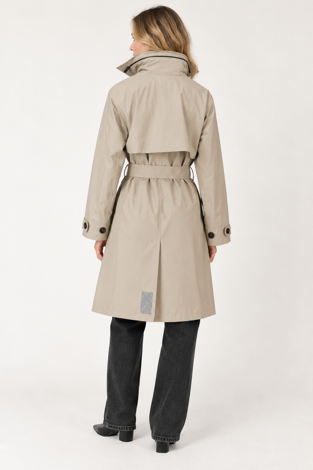 A woman shown from the back wearing a beige trench coat with a belted waist, paired with dark gray jeans and black shoes, against a plain white studio background.