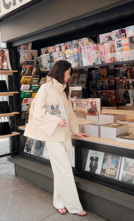 A woman wearing a cream-colored high-neck long-sleeve top and matching wide-leg trousers stands in front of a book and magazine shop window, holding a magazine.