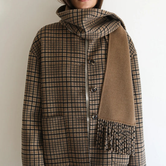 A woman wearing a brown and beige plaid coat with a matching scarf draped over one shoulder, styled with dark sunglasses and black pants against a plain white background.