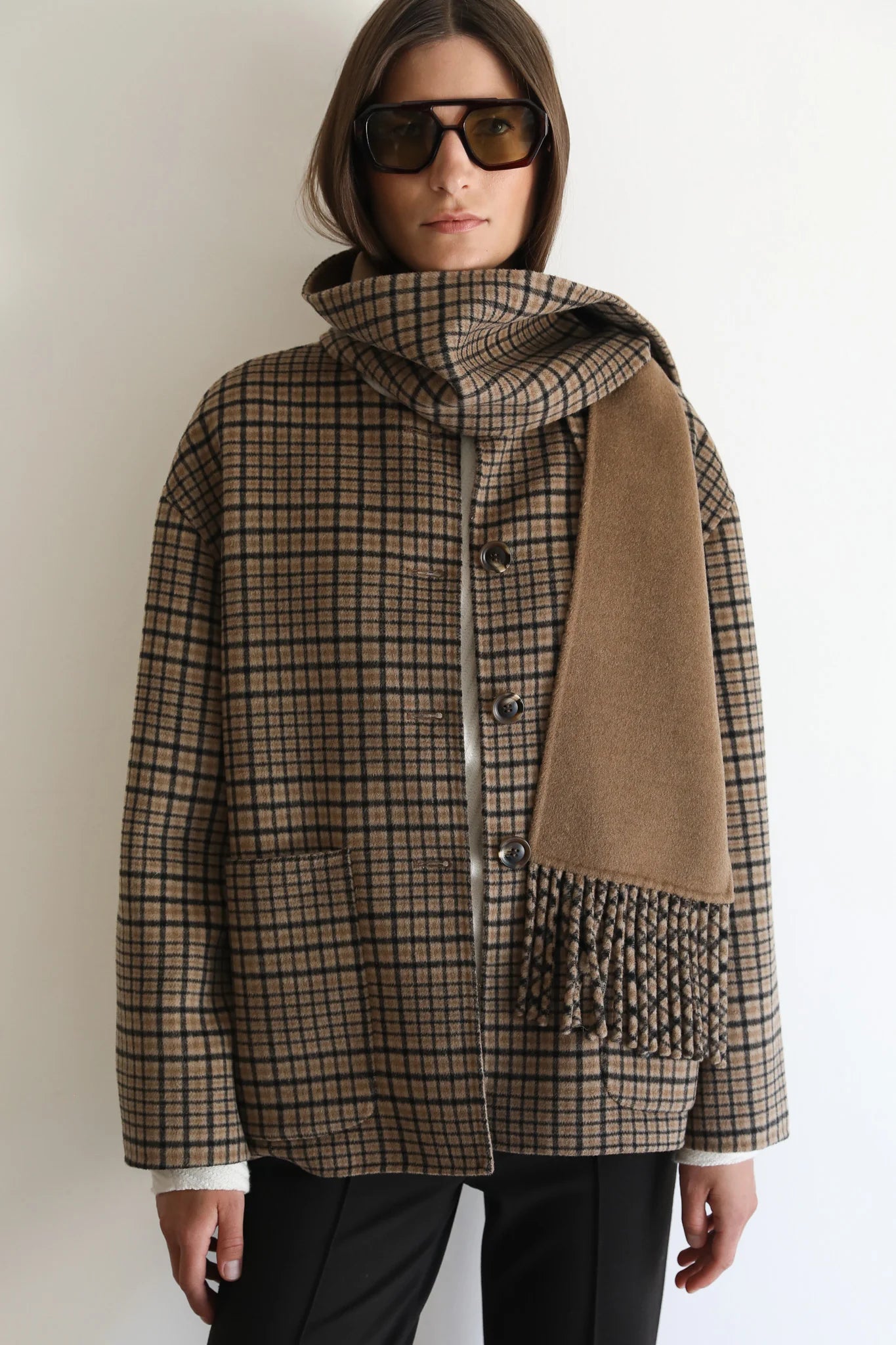 A woman wearing a brown and beige plaid coat with a matching scarf draped over one shoulder, styled with dark sunglasses and black pants against a plain white background.