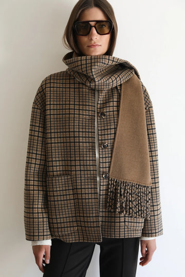 A woman wearing a brown and beige plaid coat with a matching scarf draped over one shoulder, styled with dark sunglasses and black pants against a plain white background.