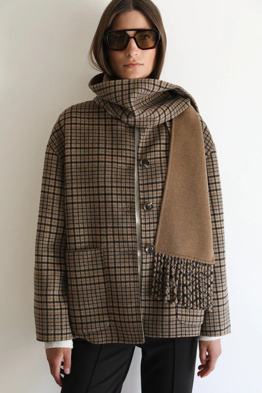 A woman wearing a brown and beige plaid coat with a matching scarf draped over one shoulder, styled with dark sunglasses and black pants against a plain white background.