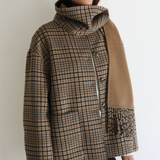 A woman wearing a brown and beige plaid cowl-neck jacket with a tan suede-like panel on the front, styled with black pants and reflective sunglasses against a plain white background.