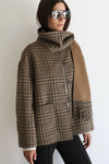 A woman wearing a brown and beige plaid cowl-neck jacket with a tan suede-like panel on the front, styled with black pants and reflective sunglasses against a plain white background.