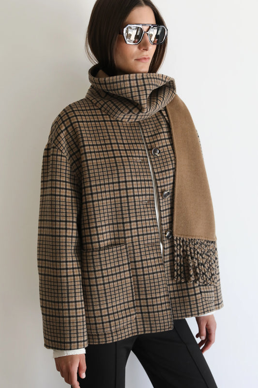 A woman wearing a brown and beige plaid cowl-neck jacket with a tan suede-like panel on the front, styled with black pants and reflective sunglasses against a plain white background.