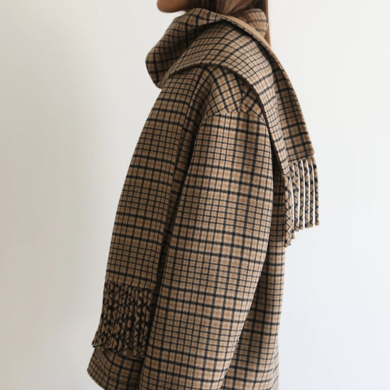 A woman shown in profile wearing a beige and brown plaid wrap coat with a matching fringed scarf, set against a plain white background.