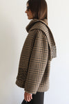A woman shown in profile wearing a beige and brown plaid wrap coat with a matching fringed scarf, set against a plain white background.