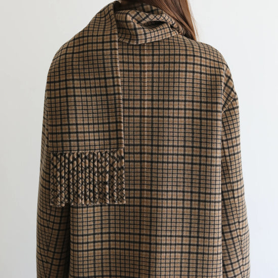 A woman viewed from behind wearing a brown and beige plaid coat with a wide collar and a fringed pocket detail on the back, set against a plain white background.