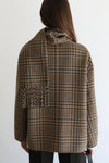 A woman viewed from behind wearing a brown and beige plaid coat with a wide collar and a fringed pocket detail on the back, set against a plain white background.