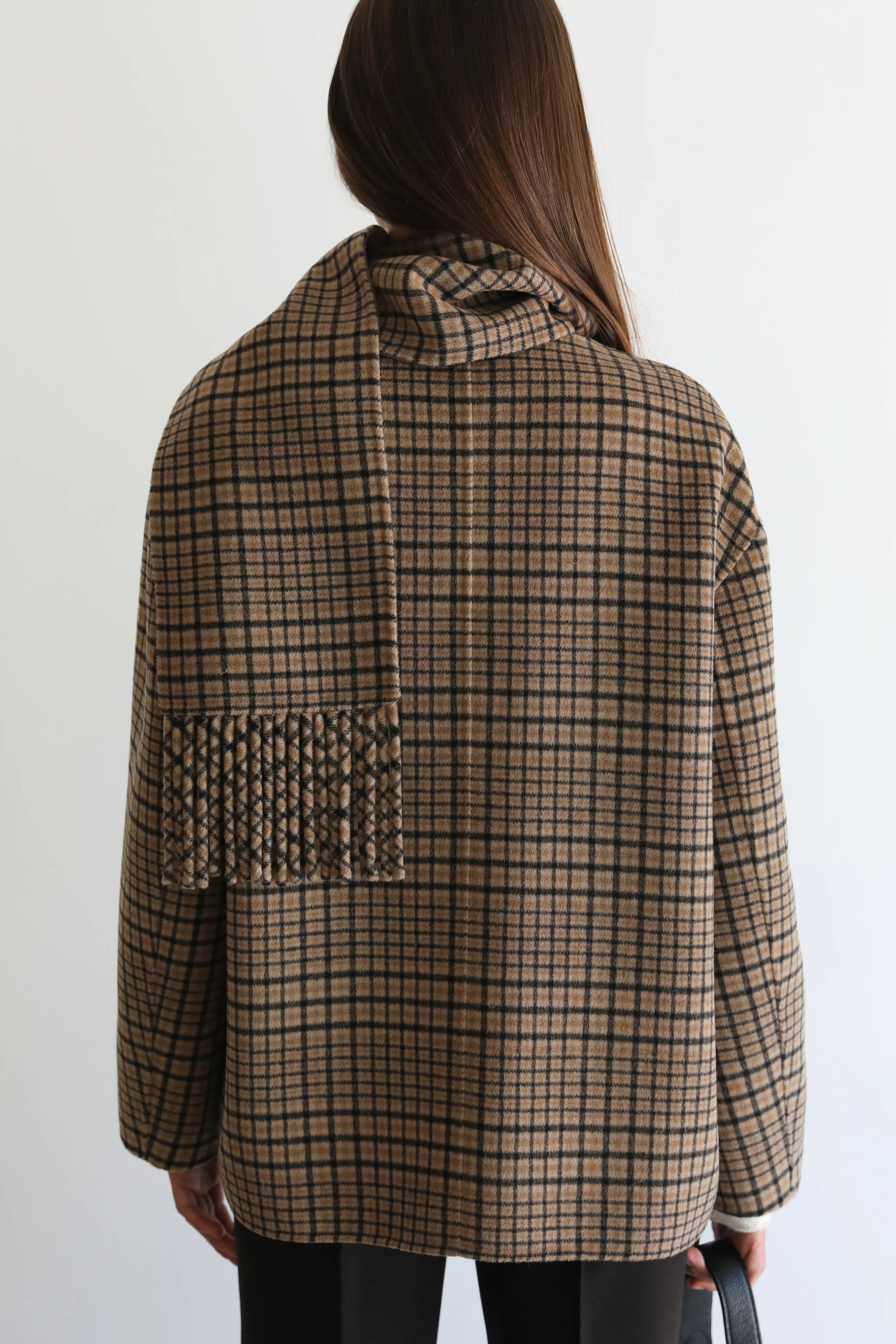 A woman viewed from behind wearing a brown and beige plaid coat with a wide collar and a fringed pocket detail on the back, set against a plain white background.