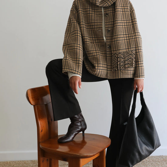 A model wearing a brown and beige plaid hooded jacket, black pants, and black boots, sitting on a wooden chair and holding a large black leather tote bag against a plain light gray wall.