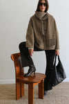 A model wearing a brown and beige plaid hooded jacket, black pants, and black boots, sitting on a wooden chair and holding a large black leather tote bag against a plain light gray wall.