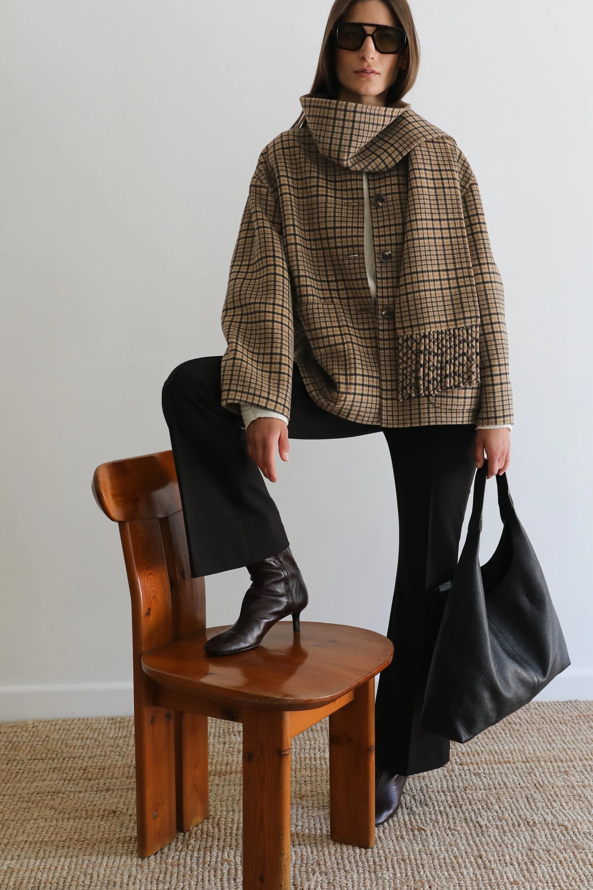 A model wearing a brown and beige plaid hooded jacket, black pants, and black boots, sitting on a wooden chair and holding a large black leather tote bag against a plain light gray wall.
