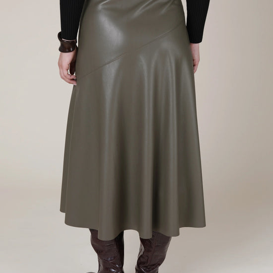 A person shown from the back wearing a high-waisted, mid-length olive green faux-leather skirt with diagonal seam details, paired with a black ribbed sweater and dark brown knee-high boots, against a solid beige studio background.