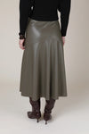 A person shown from the back wearing a high-waisted, mid-length olive green faux-leather skirt with diagonal seam details, paired with a black ribbed sweater and dark brown knee-high boots, against a solid beige studio background.