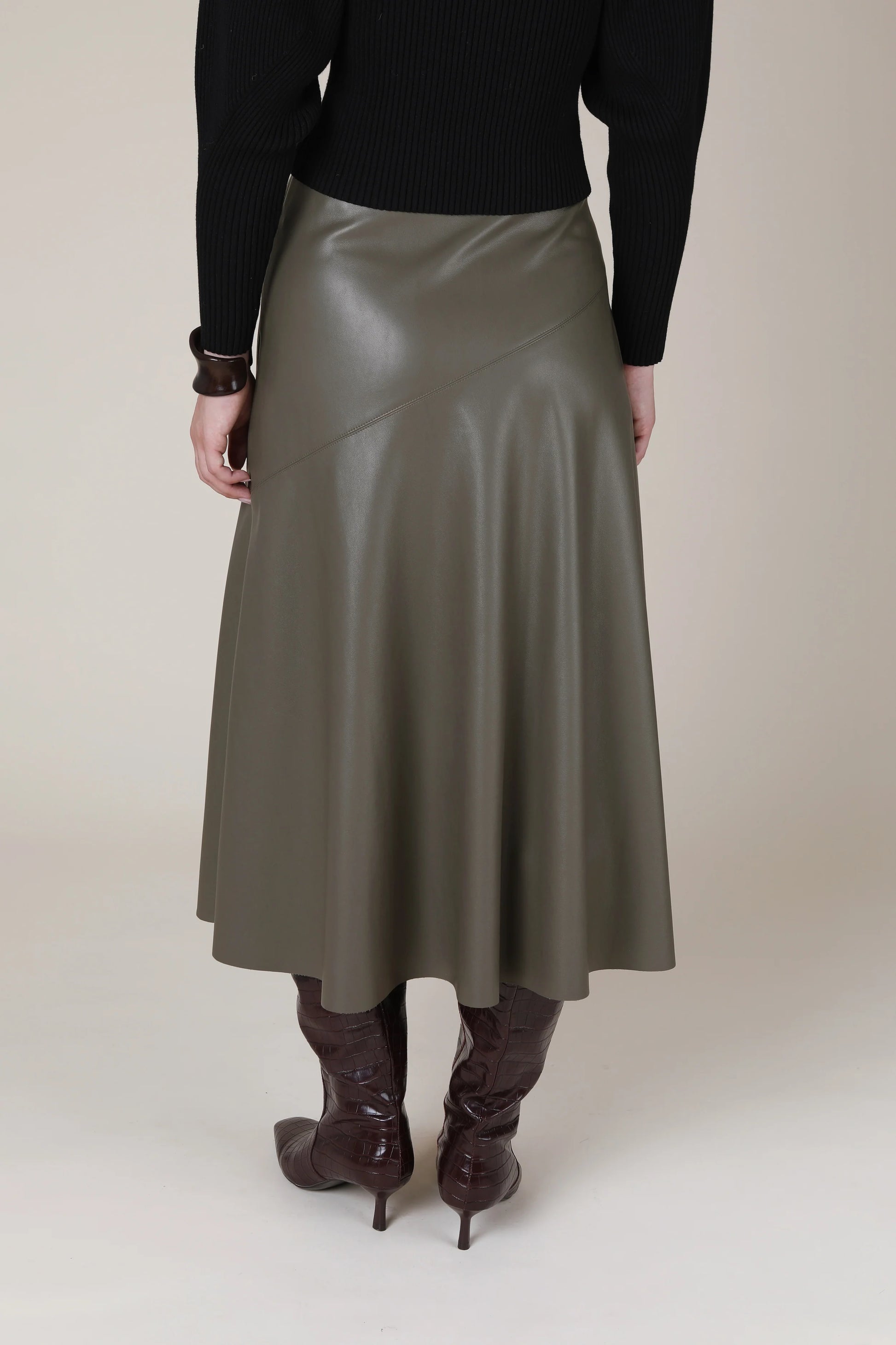A person shown from the back wearing a high-waisted, mid-length olive green faux-leather skirt with diagonal seam details, paired with a black ribbed sweater and dark brown knee-high boots, against a solid beige studio background.