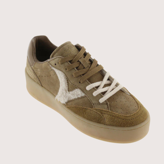 A low-top sneaker in a muted taupe-brown suede and leather mix, featuring a contrasting white fuzzy side panel and white laces, set against a solid light beige background.