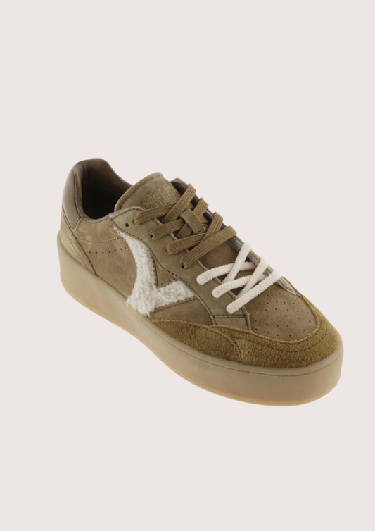 A low-top sneaker in a muted taupe-brown suede and leather mix, featuring a contrasting white fuzzy side panel and white laces, set against a solid light beige background.