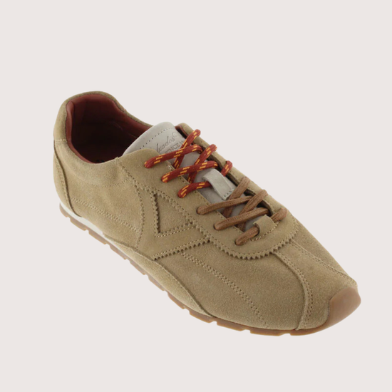 A single low-top casual sneaker in a light tan suede-like material, featuring brown laces and a brown outsole, shown on a solid light beige background.