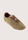 A single low-top casual sneaker in a light tan suede-like material, featuring brown laces and a brown outsole, shown on a solid light beige background.