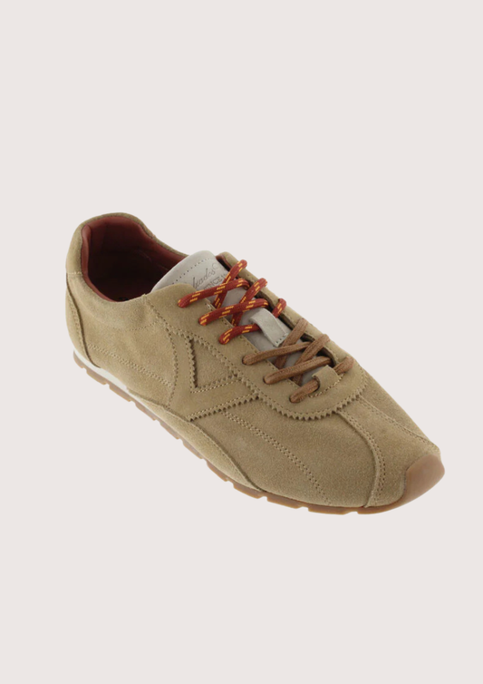 A single low-top casual sneaker in a light tan suede-like material, featuring brown laces and a brown outsole, shown on a solid light beige background.