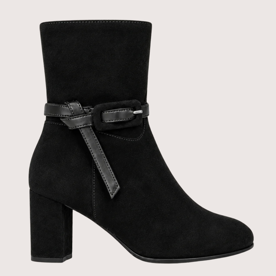A black suede ankle boot with a block heel and a decorative black leather strap with a silver buckle around the ankle, shown against a light gray background.