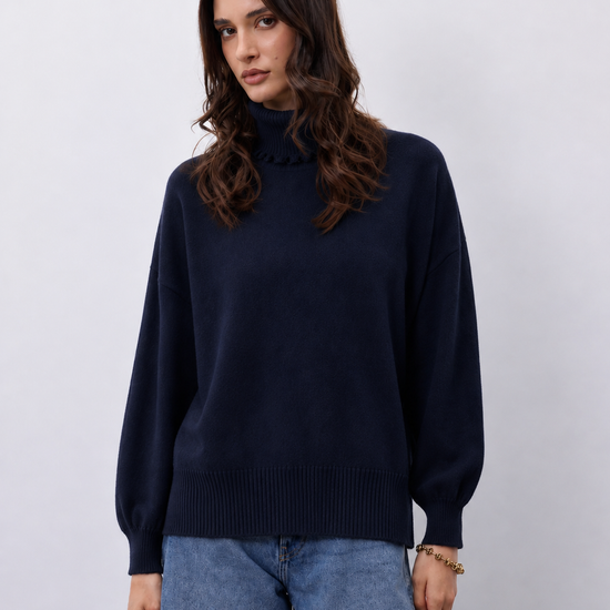 A woman wearing a dark navy blue knit mock-neck sweater and light blue jeans, standing against a light gray studio background.