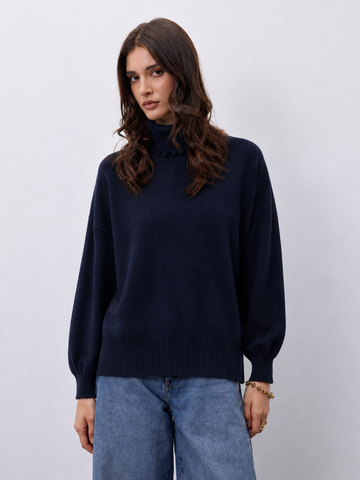 A woman wearing a dark navy blue knit mock-neck sweater and light blue jeans, standing against a light gray studio background.