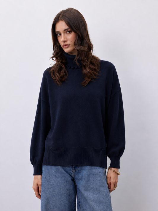 A woman wearing a dark navy blue knit mock-neck sweater and light blue jeans, standing against a light gray studio background.