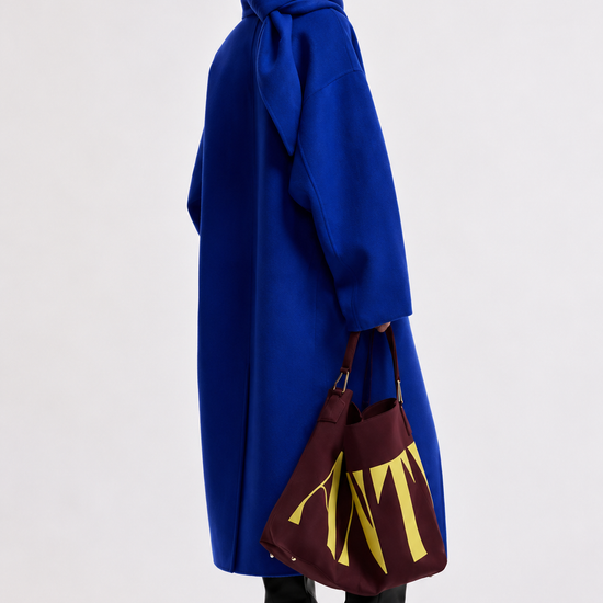 A model is shown from the back wearing a voluminous royal blue coat with a large bow at the collar, holding a burgundy tote bag with yellow lettering, set against a plain light gray studio background.