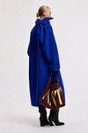 A model is shown from the back wearing a voluminous royal blue coat with a large bow at the collar, holding a burgundy tote bag with yellow lettering, set against a plain light gray studio background.
