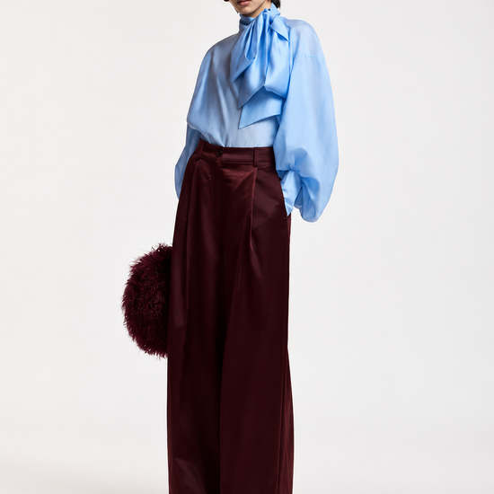 A model stands against a plain light background wearing a light blue high-neck blouse with a large bow at the collar and voluminous sleeves, paired with high-waisted wide-leg burgundy trousers. She holds a dark burgundy fuzzy accessory.
