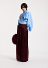 A model stands against a plain light background wearing a light blue high-neck blouse with a large bow at the collar and voluminous sleeves, paired with high-waisted wide-leg burgundy trousers. She holds a dark burgundy fuzzy accessory.