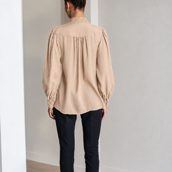 A woman viewed from behind wearing a beige long-sleeve blouse with gathered puff sleeves and a high gathered neckline, paired with black pants and black boots, standing in a minimalist room with white walls and light wood flooring.