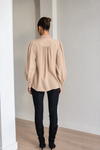 A woman viewed from behind wearing a beige long-sleeve blouse with gathered puff sleeves and a high gathered neckline, paired with black pants and black boots, standing in a minimalist room with white walls and light wood flooring.