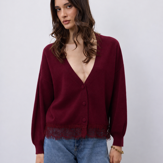 A woman wearing a deep burgundy cardigan with a V-neck, button front, and fringe trim, paired with blue jeans, against a plain light gray background.