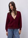 A woman wearing a deep burgundy cardigan with a V-neck, button front, and fringe trim, paired with blue jeans, against a plain light gray background.