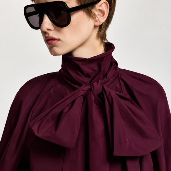 A model with short brown hair wearing oversized black sunglasses and a high-neck, deep burgundy blouse with a large bow at the collar, photographed against a light gray studio background.