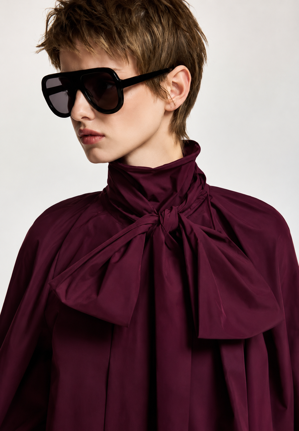 A model with short brown hair wearing oversized black sunglasses and a high-neck, deep burgundy blouse with a large bow at the collar, photographed against a light gray studio background.