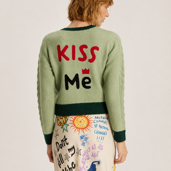 A model shown from the back wearing a sage green crewneck sweater with 'KISS Me' written on the back in red and black, paired with a cream-colored skirt featuring colorful doodles and text.