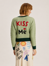 A model shown from the back wearing a sage green crewneck sweater with 'KISS Me' written on the back in red and black, paired with a cream-colored skirt featuring colorful doodles and text.