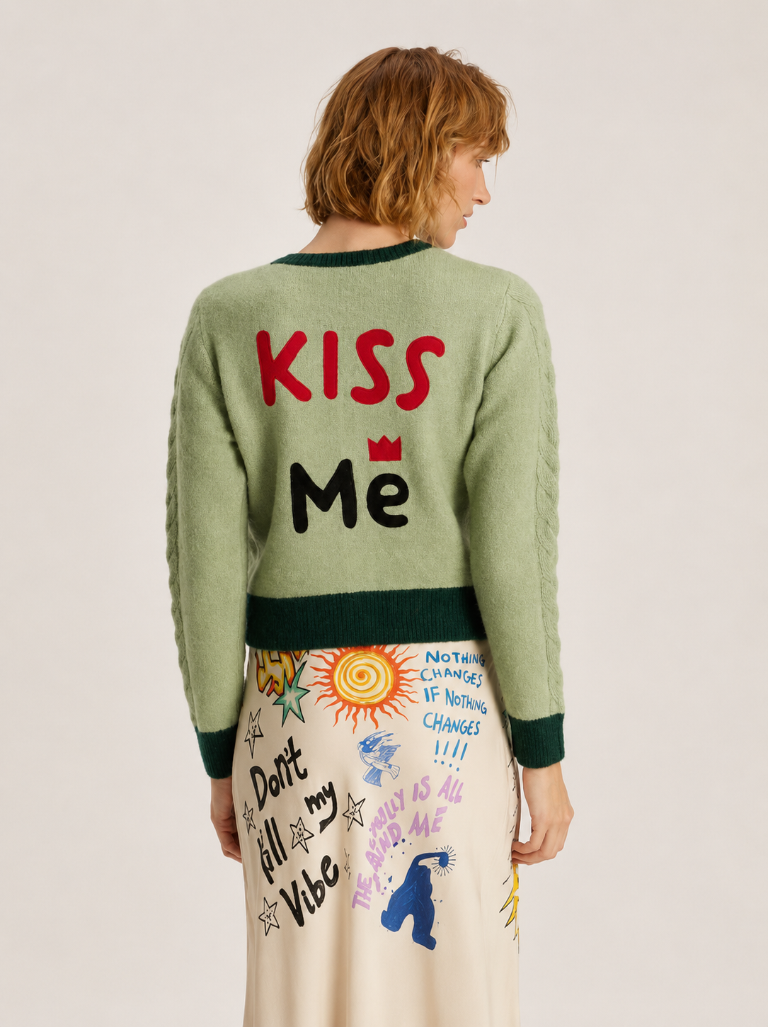 A model shown from the back wearing a sage green crewneck sweater with 'KISS Me' written on the back in red and black, paired with a cream-colored skirt featuring colorful doodles and text.