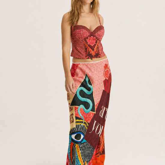 A woman modeling a two-piece outfit consisting of a red patterned strapless crop top and a long, wide-leg skirt with a colorful abstract print in pink, red, blue, and black, set against a plain light gray studio background.