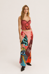 A woman modeling a two-piece outfit consisting of a red patterned strapless crop top and a long, wide-leg skirt with a colorful abstract print in pink, red, blue, and black, set against a plain light gray studio background.