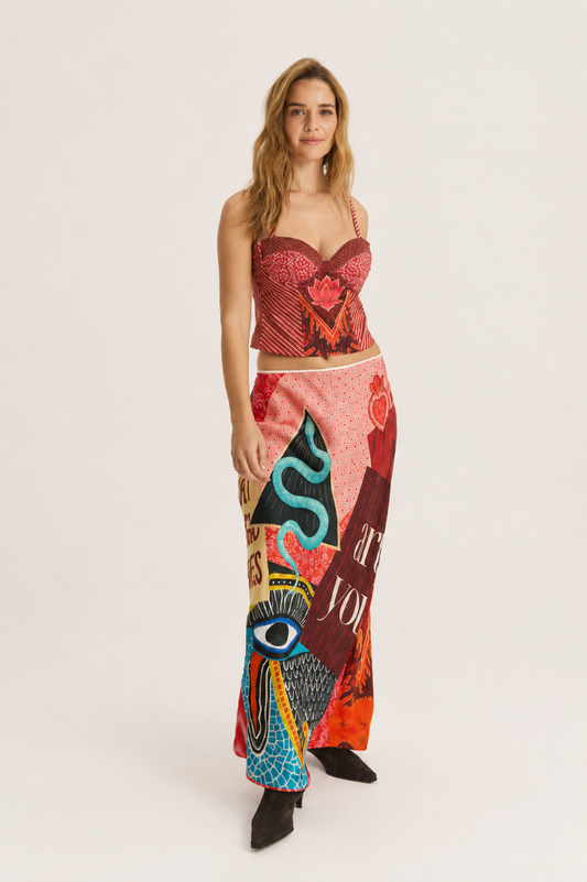A woman modeling a two-piece outfit consisting of a red patterned strapless crop top and a long, wide-leg skirt with a colorful abstract print in pink, red, blue, and black, set against a plain light gray studio background.