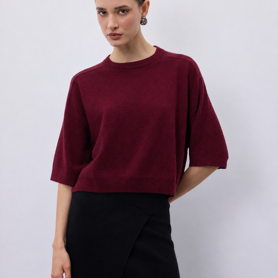 A woman modeling a short-sleeve crewneck sweater in deep burgundy, paired with a black skirt, against a plain light gray studio background.