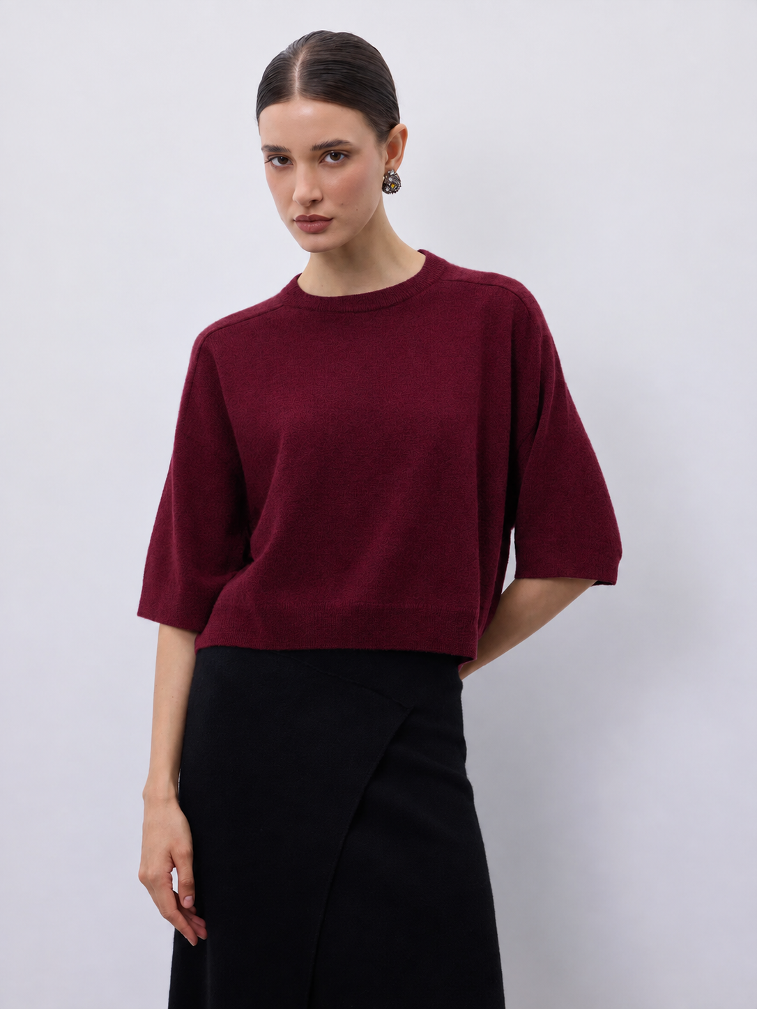 A woman modeling a short-sleeve crewneck sweater in deep burgundy, paired with a black skirt, against a plain light gray studio background.